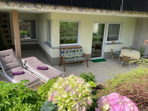 Sonnige Terrasse mit Liegen, Tisch und Stühlen, umgeben von Pflanzen und Blumen.